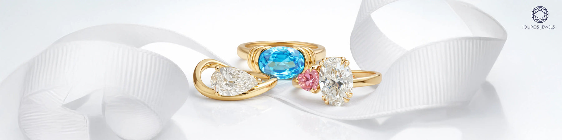 Three unique engagement rings in yellow gold featuring oval blue topaz east-west ring, oval diamond with pink diamond toi et moi ring and pear diamond wave ring by Ouros Jewels