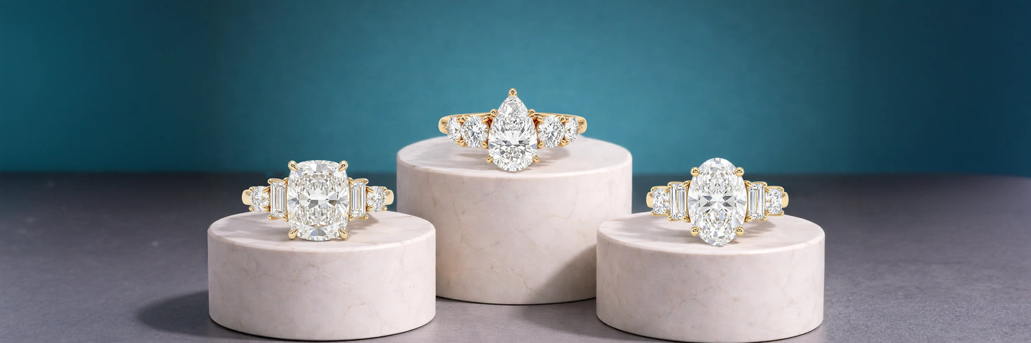 Three five-stone lab-grown diamond engagement rings in yellow gold displayed on white pedestals — cushion cut with baguette side stones, pear cut with round side stones, and oval cut with baguette side stones