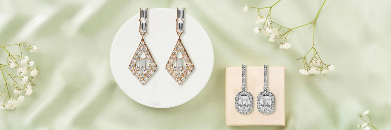 Three pairs of lab-grown diamond drop and dangle earrings on gold stands — round and pear three-stone drop earrings, bezel-set pear cut drop earrings, and marquise and pear cascade dangle earrings in white gold on marble surface