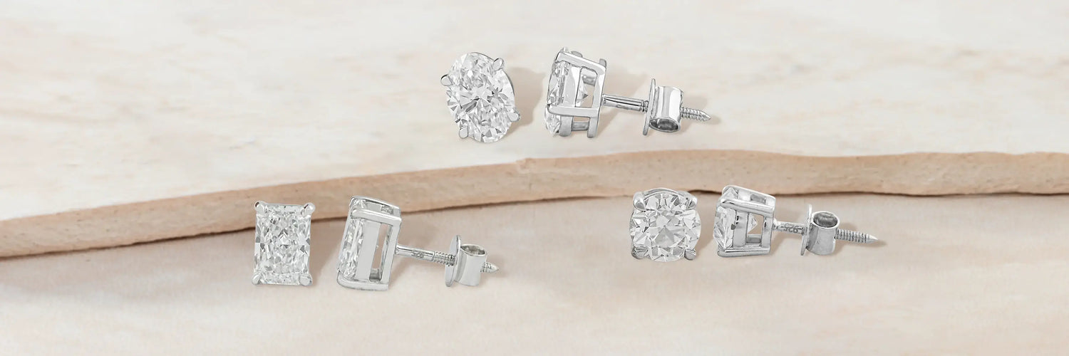 Lab-grown diamond stud earrings — heart cut halo studs in white gold, mixed emerald and heart cut two-stone studs in white gold, and round brilliant floral cluster studs in yellow gold on tiered camel sand display