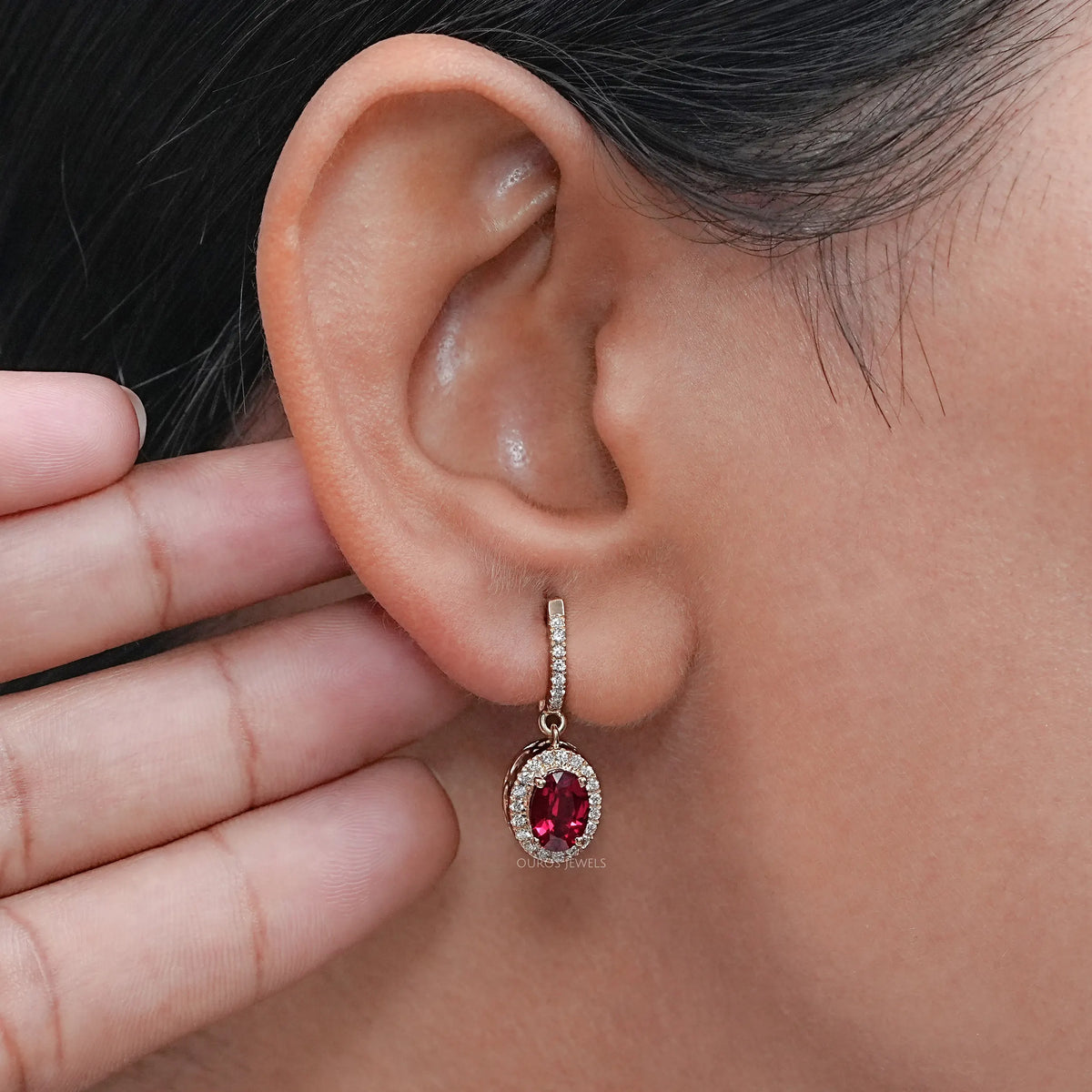 Oval cut red ruby huggie hoop earring with halo setting worn on ear